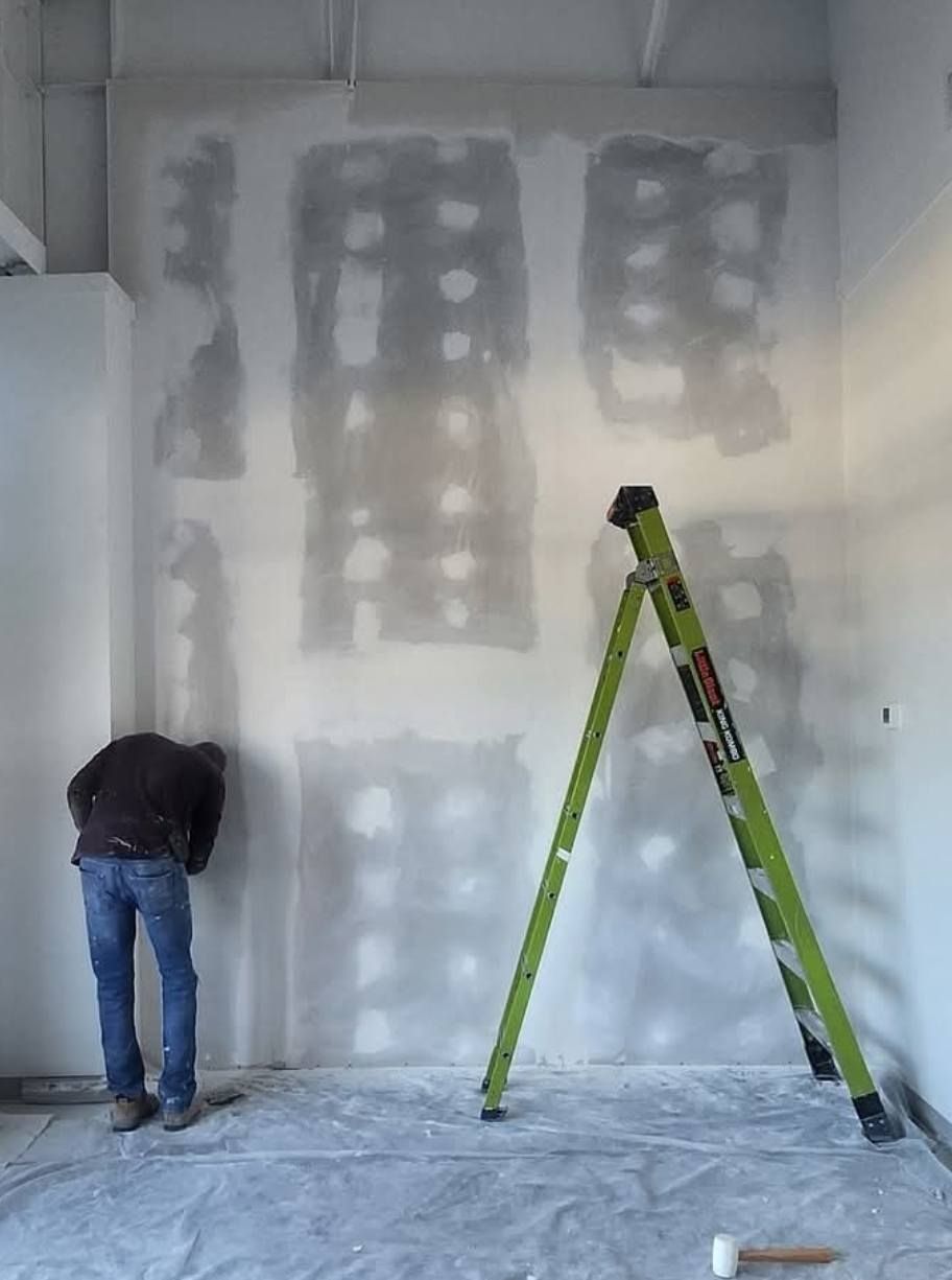 Person working on drywall, green ladder in room.