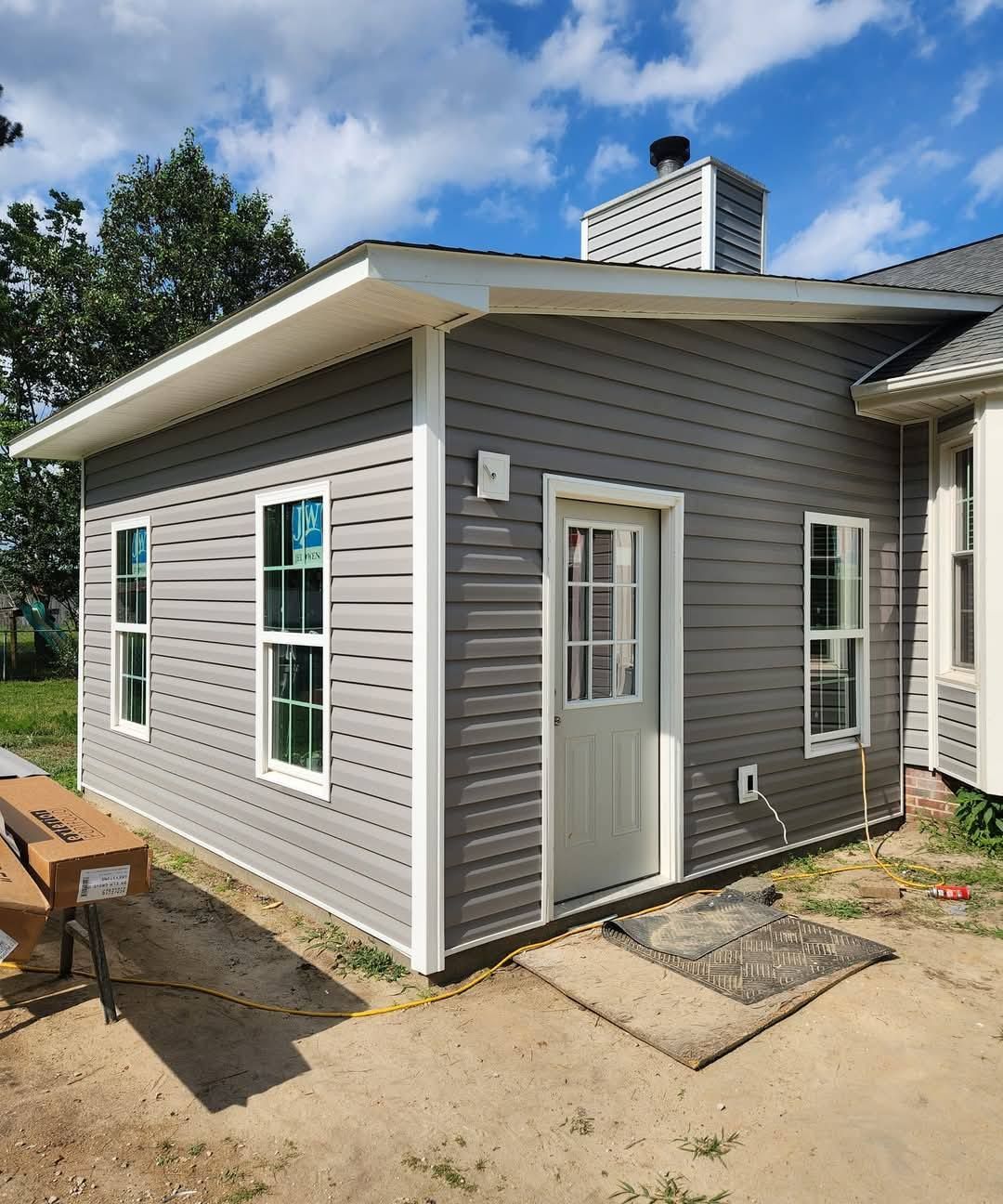 Gray-sided building with white trim, windows, and door. A chimney and the edge of a larger house are also visible.