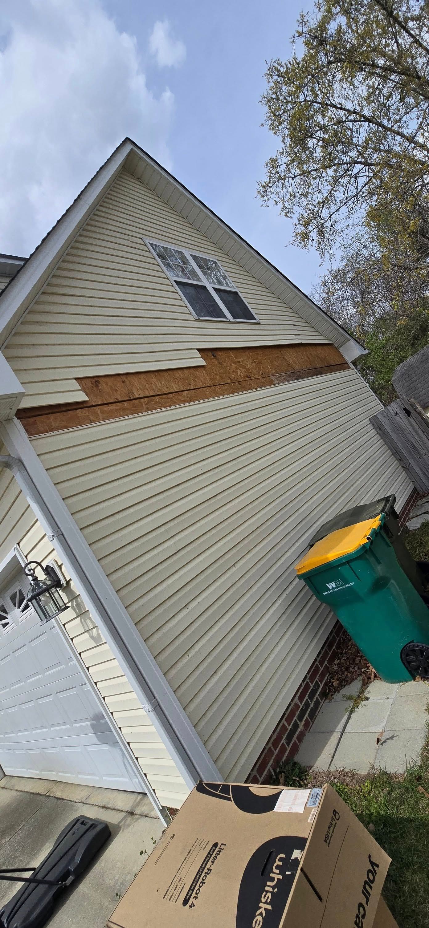 Exterior of a house with damaged siding and exposed wood, leaning. A trash bin is visible.