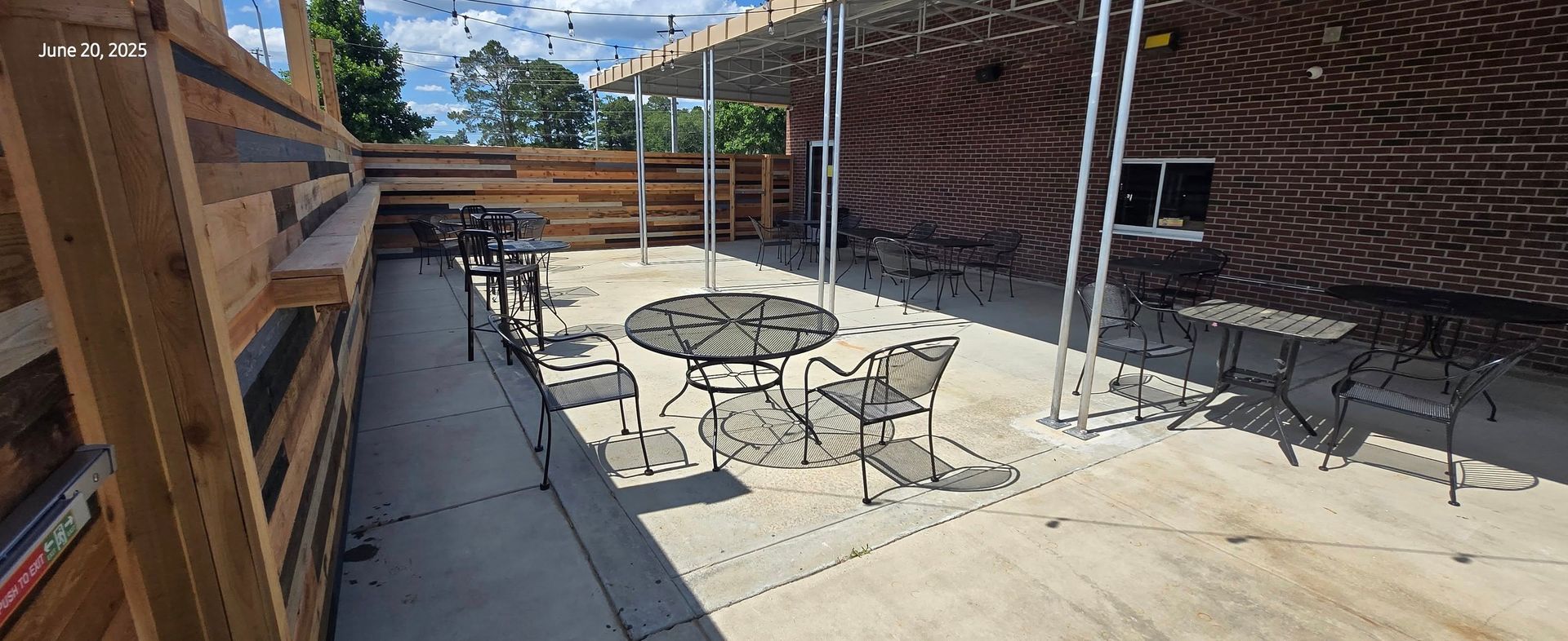 Outdoor patio with metal tables and chairs, under a shaded pergola, next to a brick building.