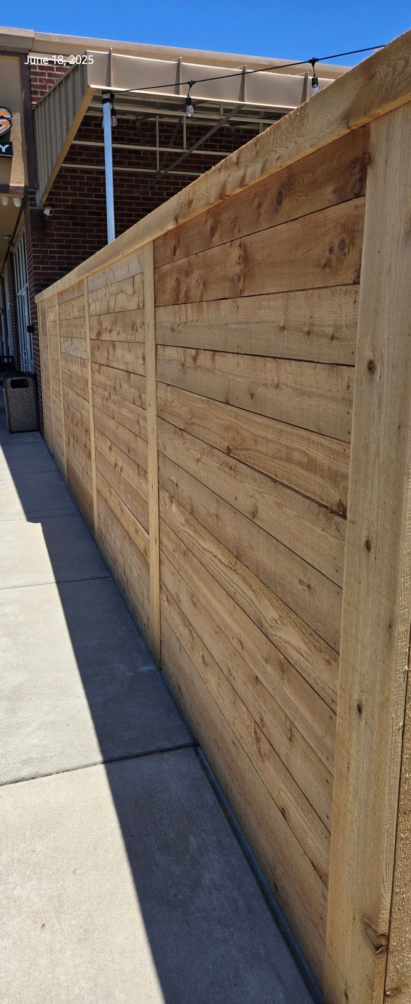 Wooden fence along a concrete walkway, with a partial view of a structure and blue sky.