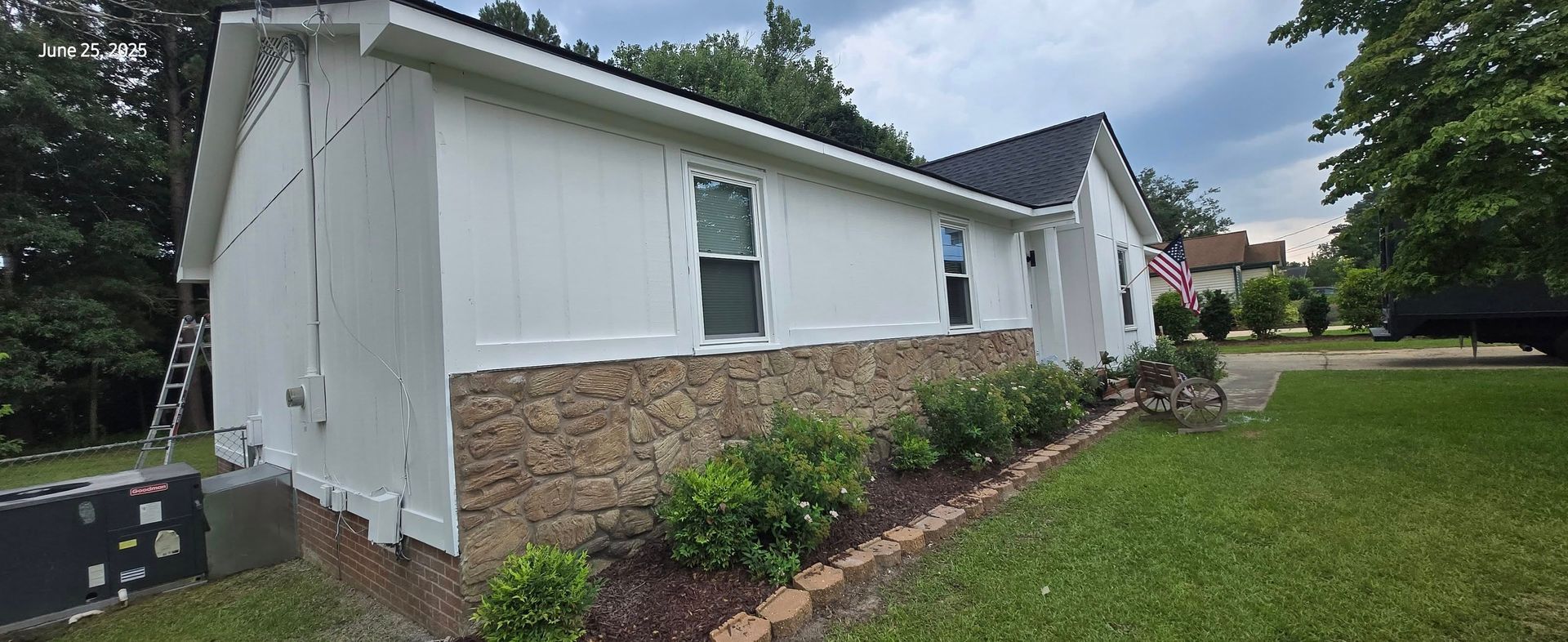 A white house with a stone base and black roof on a cloudy day. Green plants in front of the house.