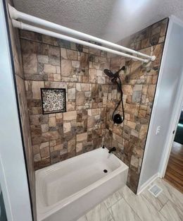 Shower with stone-look wall panels, black fixtures, and a white tub. A double shower curtain rod hangs above.