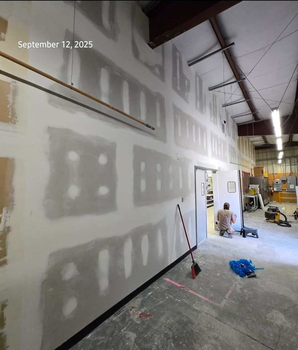 Construction in progress; drywall being installed on a wall inside a building. A person is working in the background.