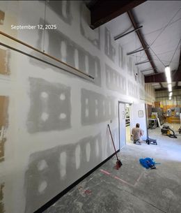 Construction in progress; drywall being installed on a wall inside a building. A person is working in the background.