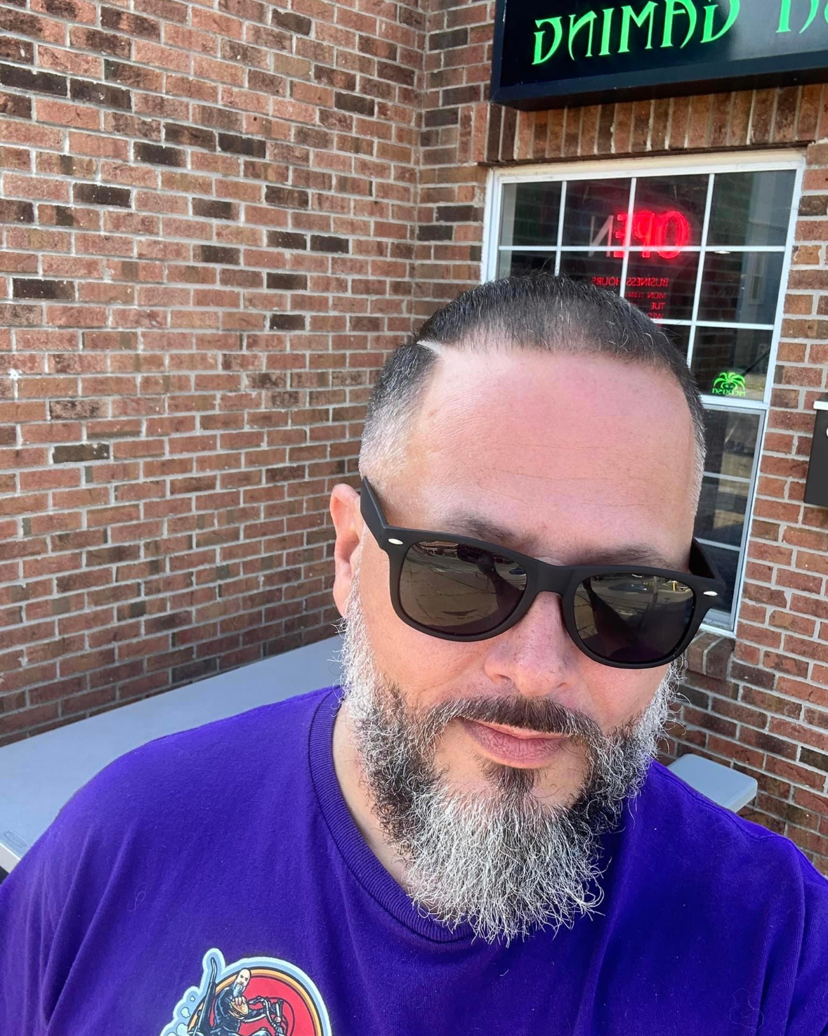 a man with a beard wearing sunglasses and a purple shirt is standing in front of a brick building .