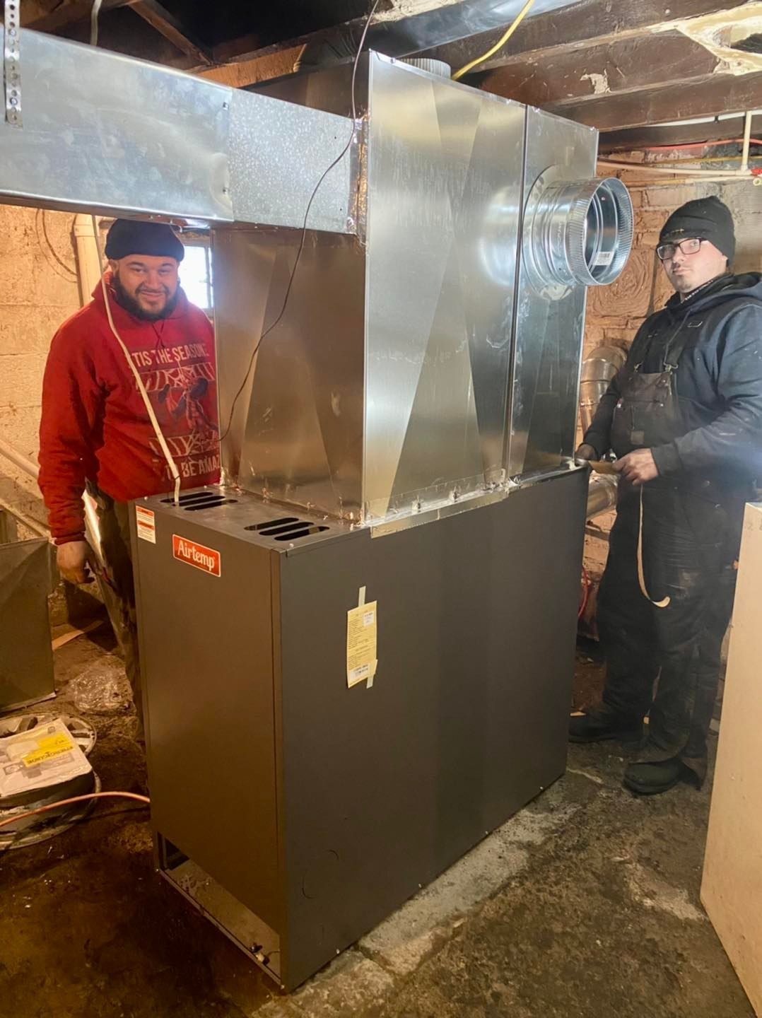 Two men are standing next to a large heater in a basement.