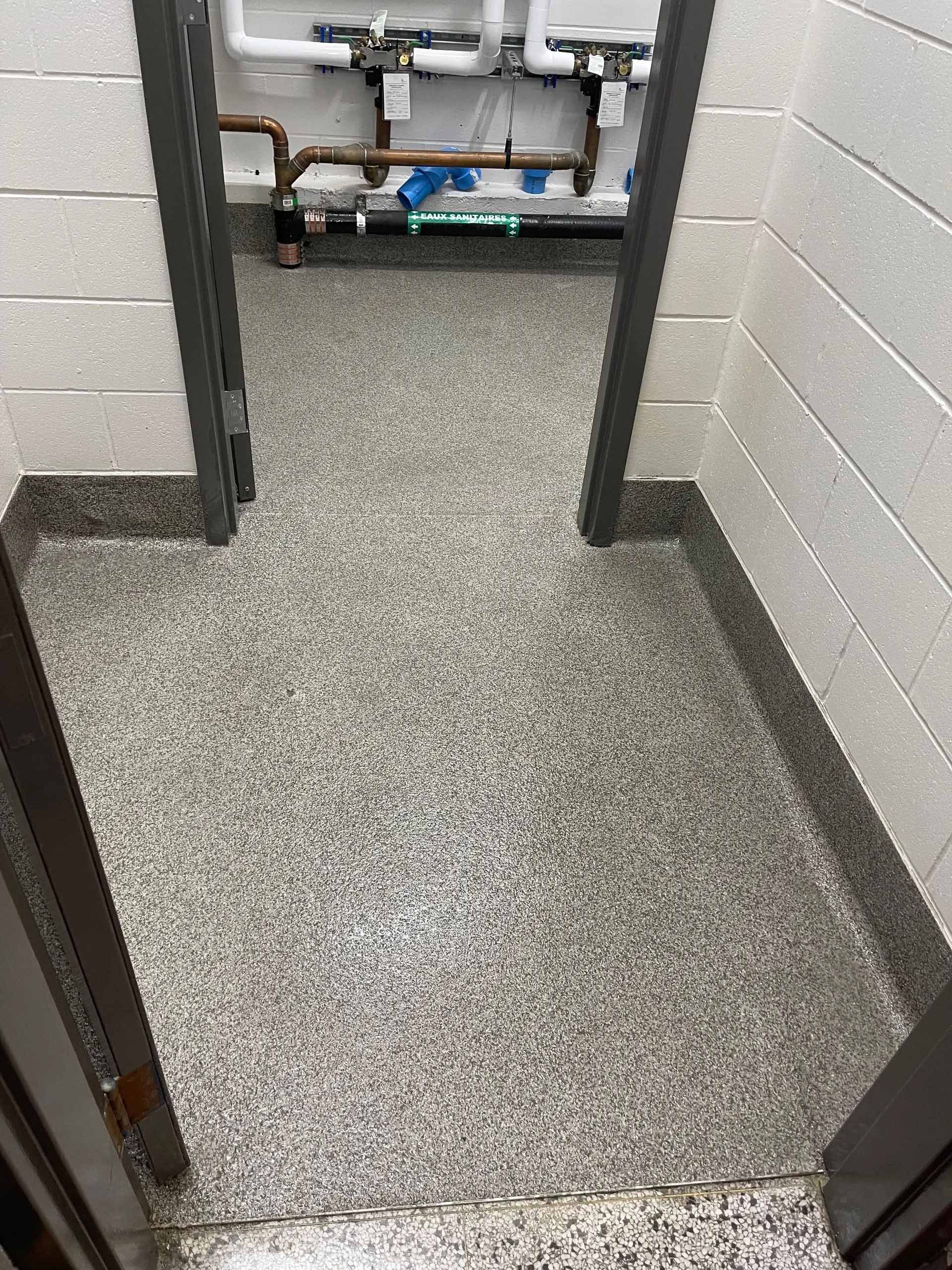 Une porte avec un sol gris moucheté et des moulures sombres. Des tuyaux sont visibles dans la pièce suivante.