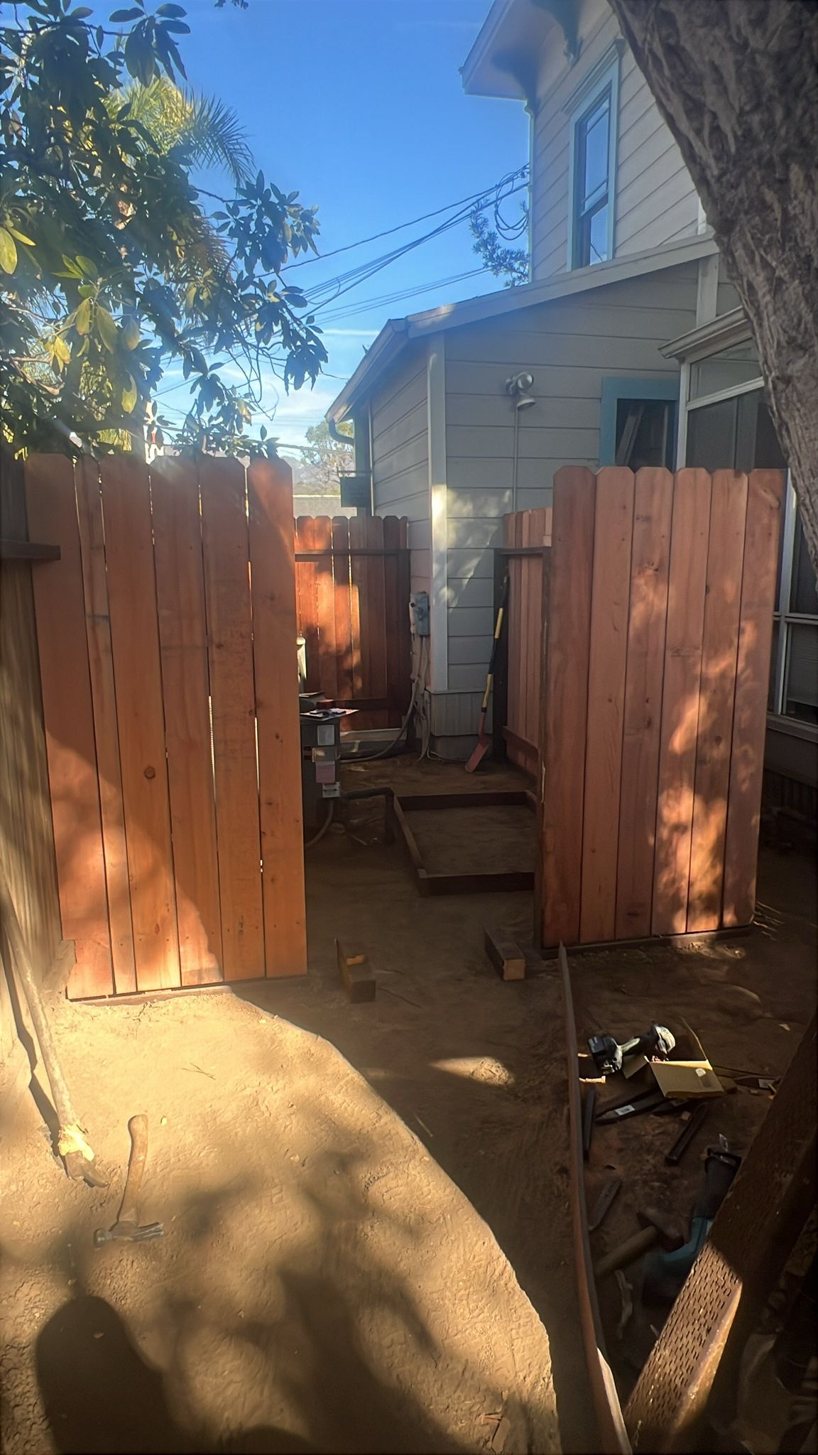 A wooden fence encloses a dirt yard with a building in the background on a sunny day.
