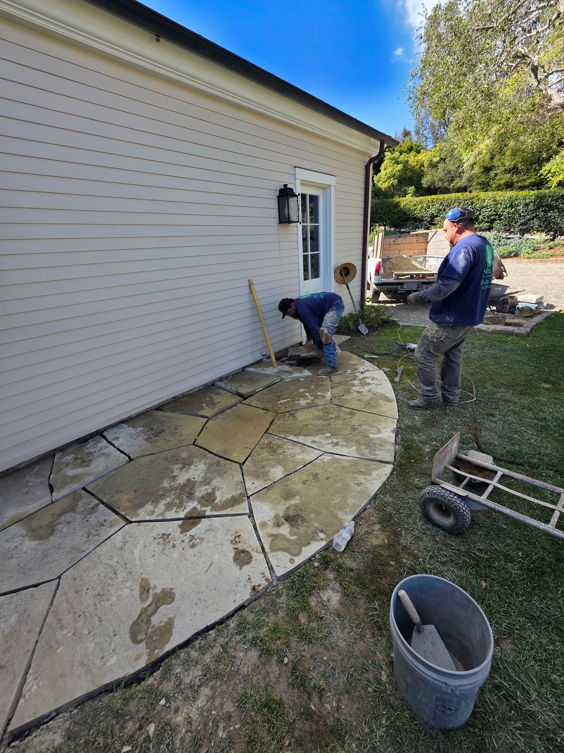 Two workers installing stone patio near a beige building. One bends over, the other stands nearby. Outdoors, sunny.