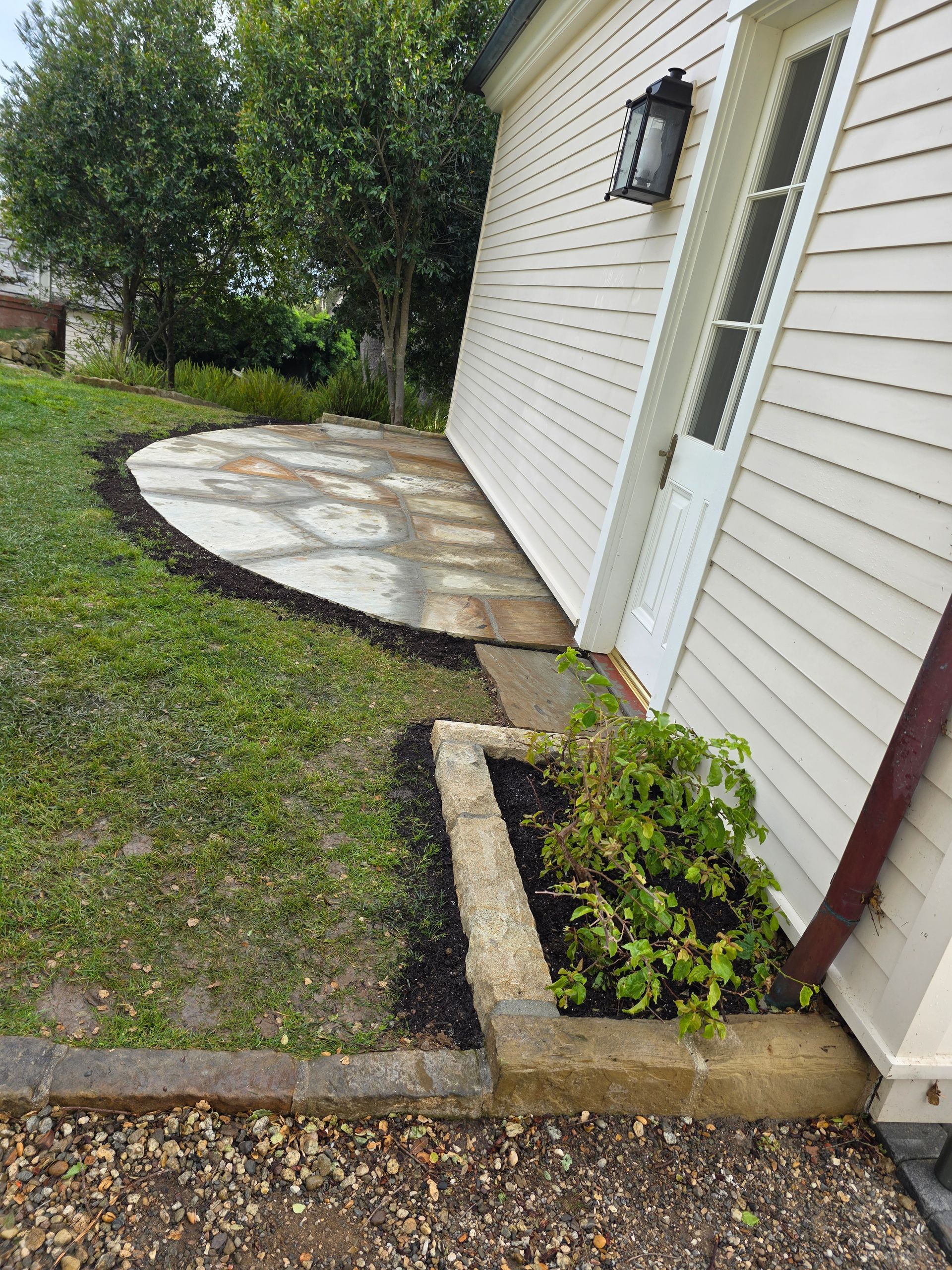 Side of a building with a small stone patio, garden bed, and doorway. Lawn, trees, and gray skies are in view.