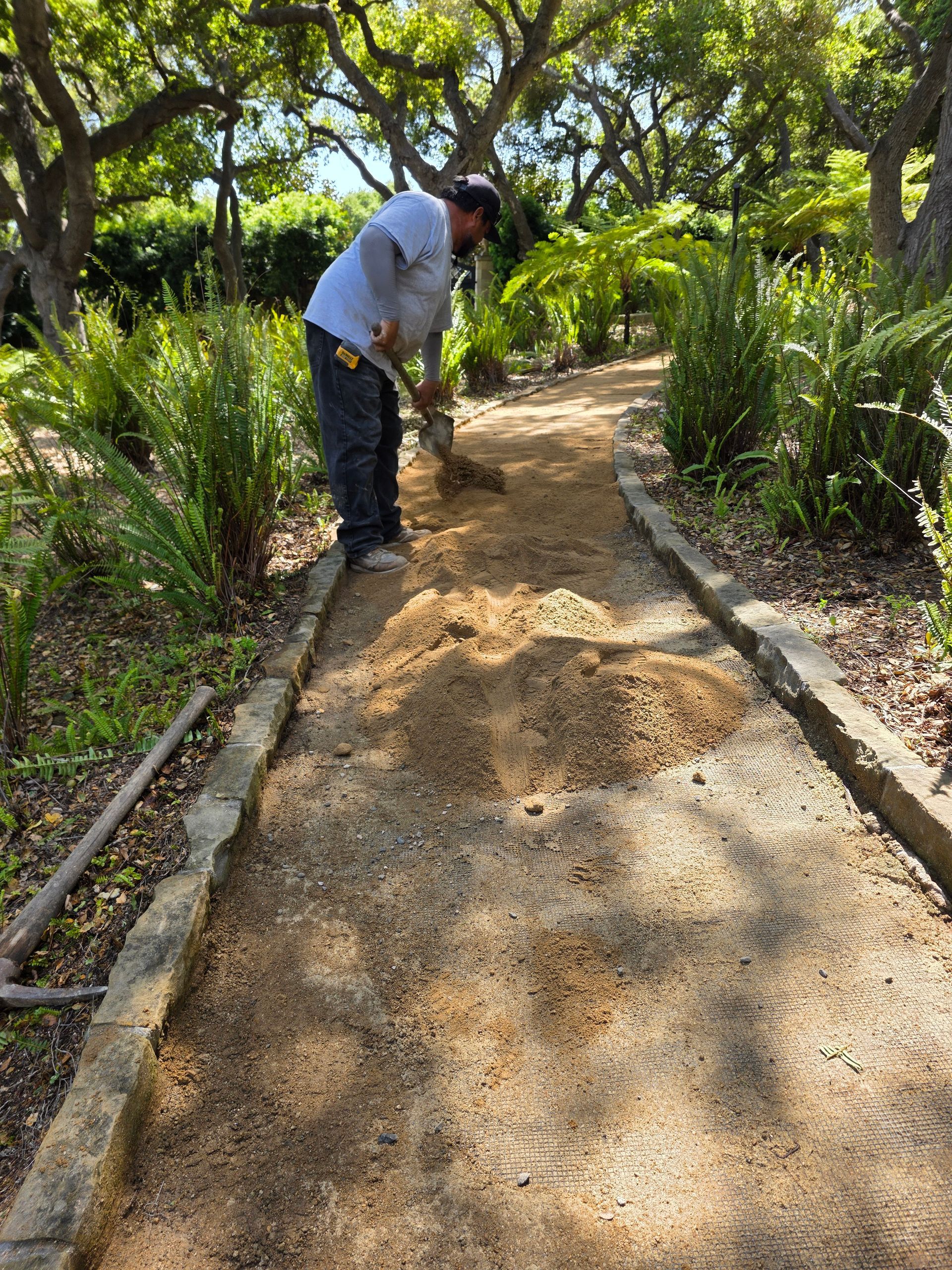 Man shoveling gravel on a garden path lined with stone edging. Lush greenery surrounds him under a sunny sky.