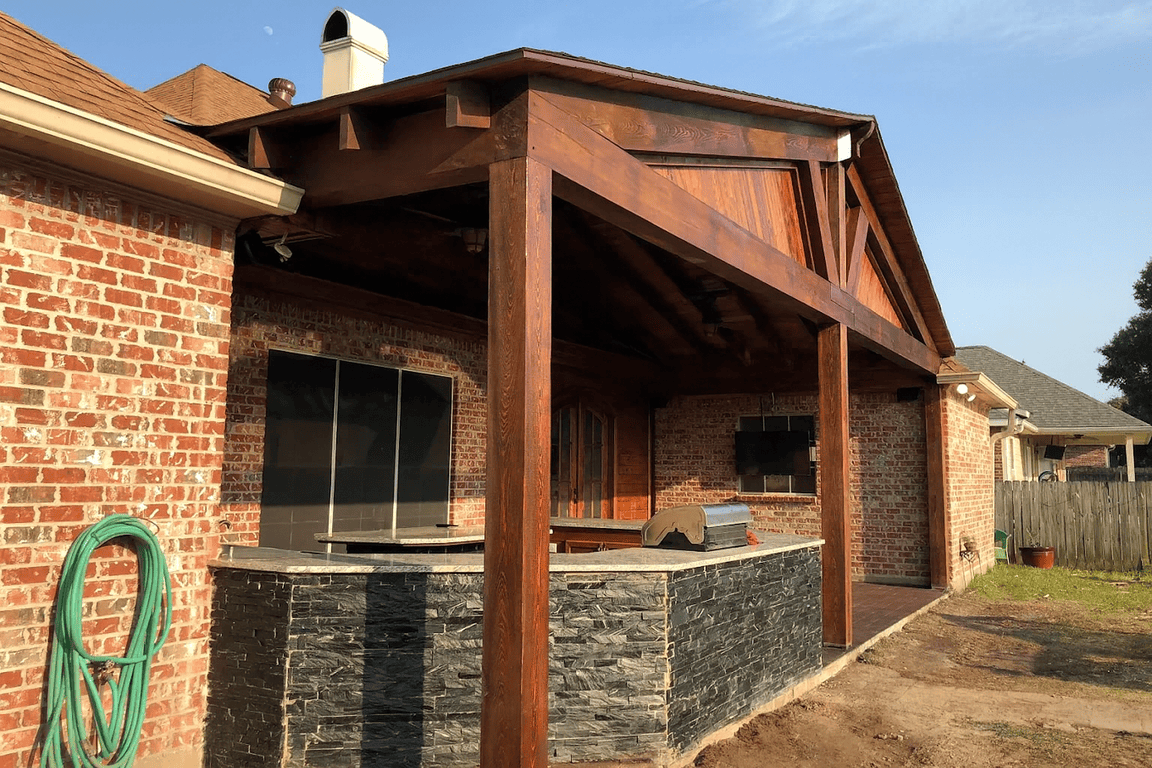 Brick house with a wooden-covered patio. Outdoor kitchen remodel in Lafayette, LA, featuring black stone.