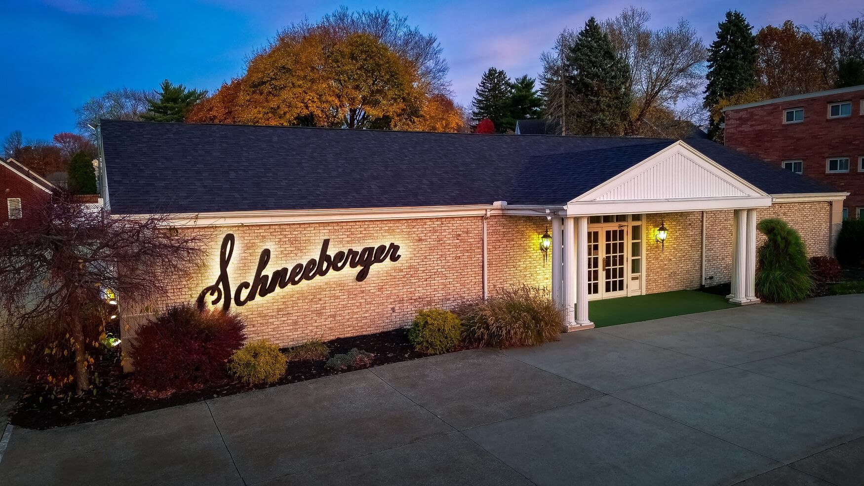 Schneeburger storefront at dusk, with lit entrance and brick exterior surrounded by trees.