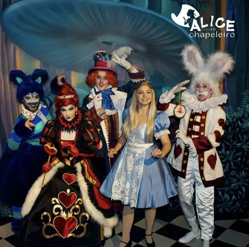 Elenco da peça "Alice no País das Maravilhas": Alice com o Chapeleiro Maluco, a Rainha de Copas, o Coelho Branco e o Gato de Cheshire. Cenário e figurinos coloridos.