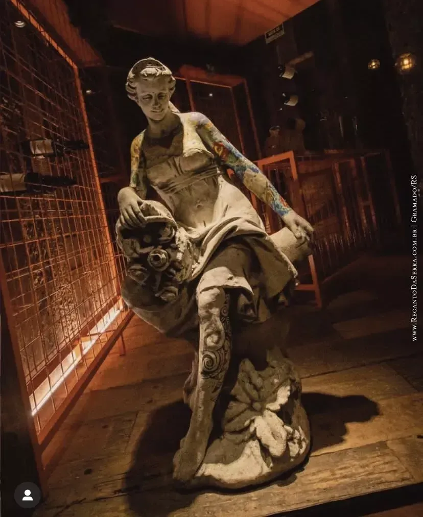 Estátua com tatuagens em sala escura com painéis de madeira. Figura feminina com tatuagens nos braços e pernas.