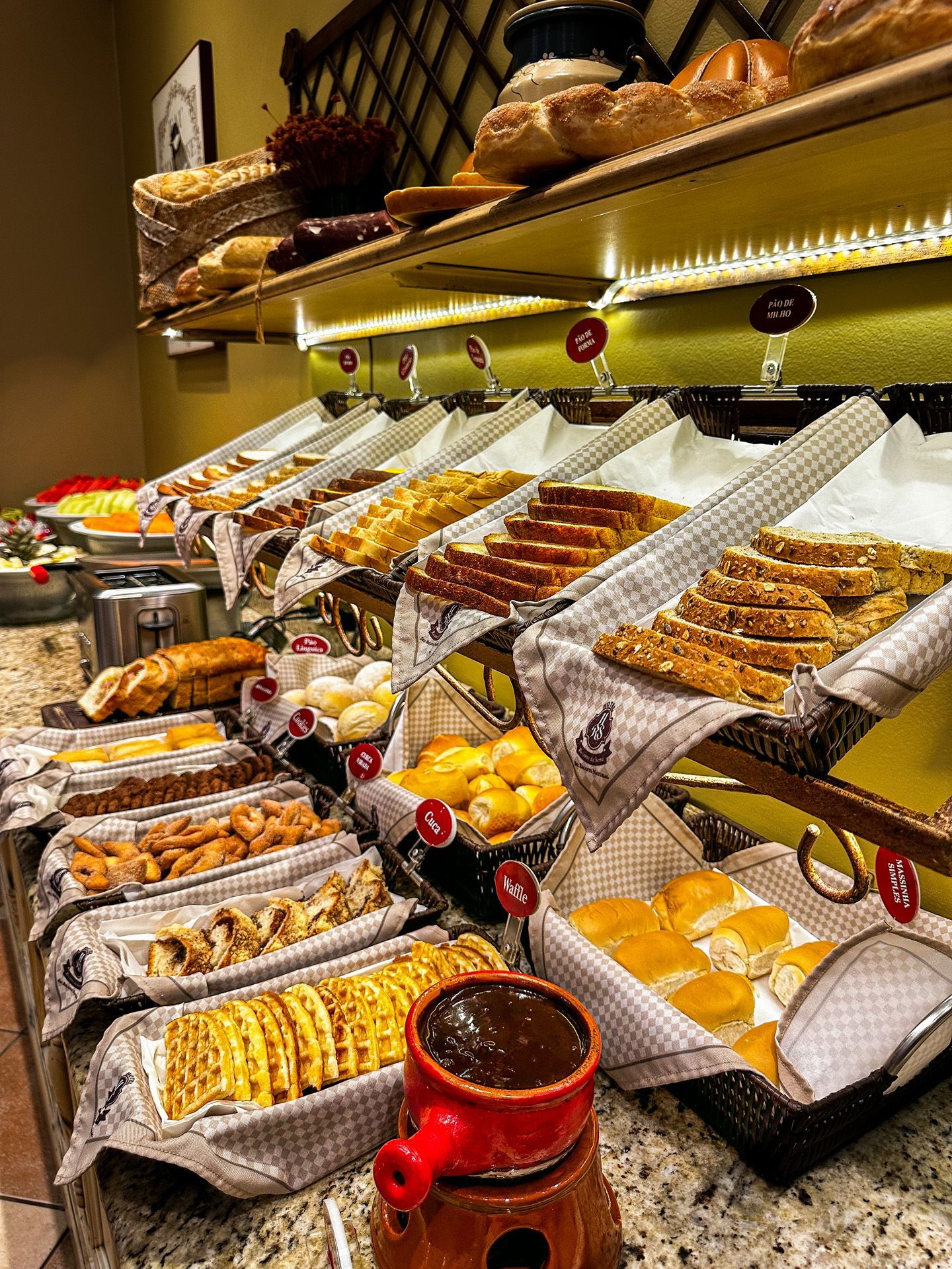 Buffet com diversos pães, doces, saladas e condimentos expostos em um balcão de madeira em um hotel.