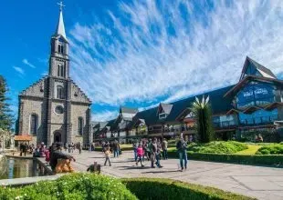 Uma igreja e lojas em Gramado, Brasil. Pessoas caminham em uma praça sob um céu azul.