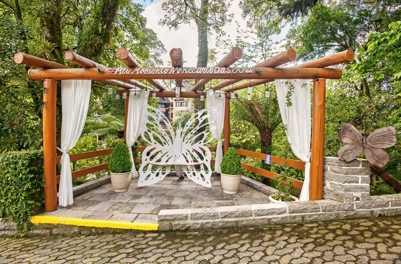 Gazebo de madeira com cortinas brancas, escultura de borboleta, vasos de plantas e caminho de pedra.