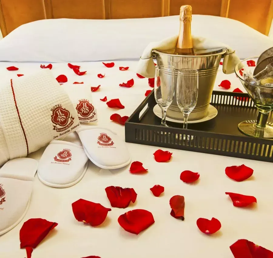 Cama decorada com pétalas de rosa, champanhe, taças de champanhe, roupão, chinelos e bandeja.