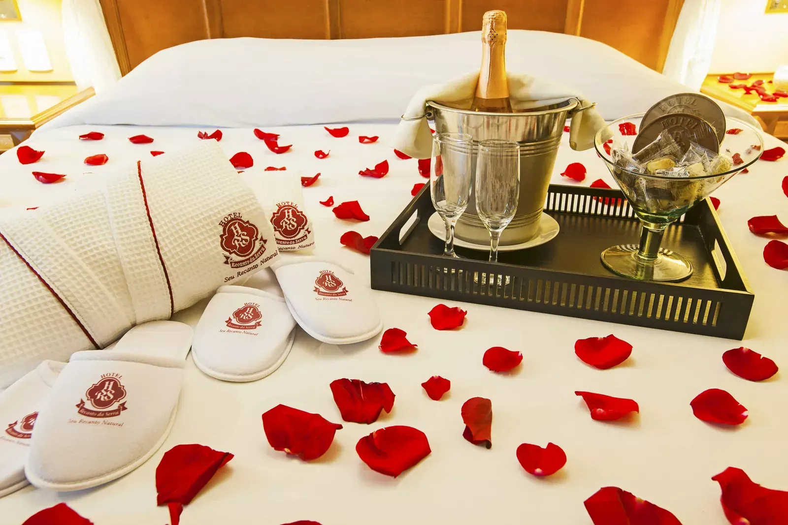 Cama decorada com pétalas de rosa, champanhe, chocolates, roupões e chinelos, um cenário romântico de hotel.
