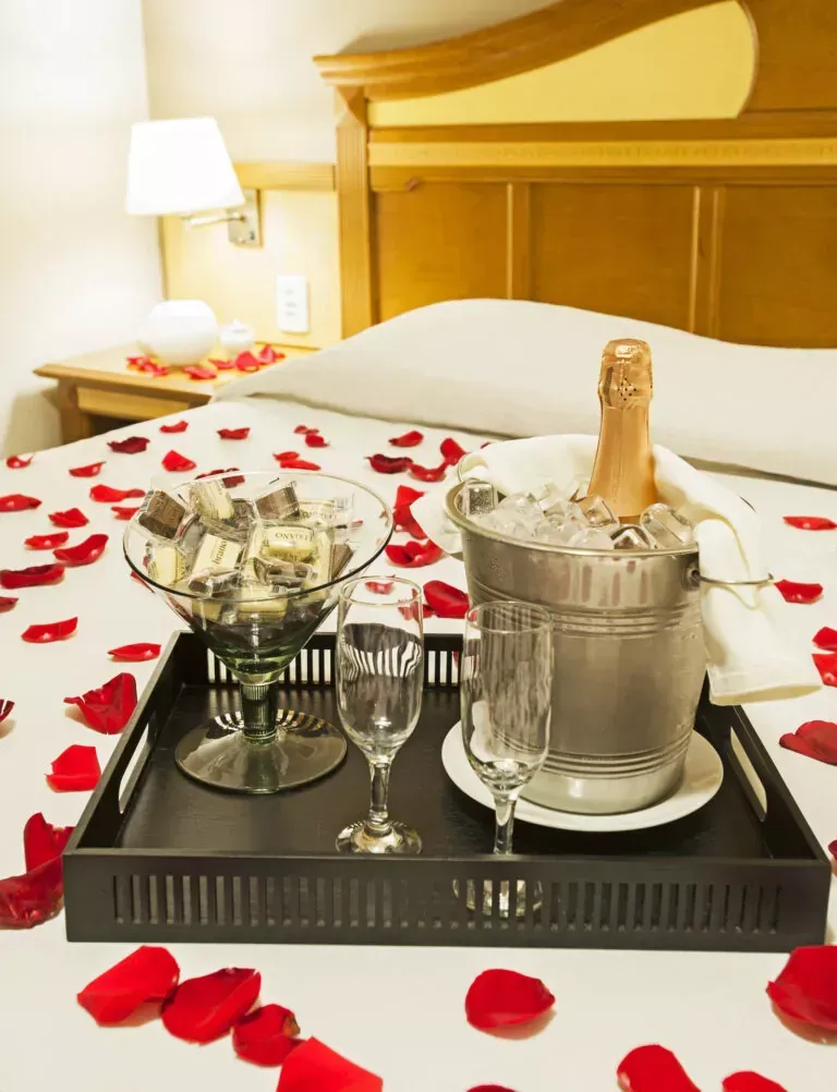 Quarto de hotel romântico com pétalas de rosa, champanhe em balde de gelo, chocolates e duas taças sobre a cama.