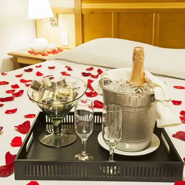 Cama com pétalas de rosa, champanhe, chocolates e duas taças em uma bandeja. Ambiente romântico.