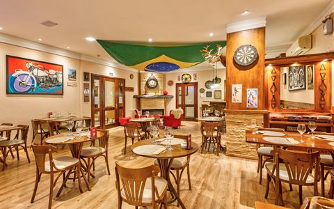 Interior de um restaurante decorado com uma bandeira brasileira, bar, mesas e poltronas vermelhas.