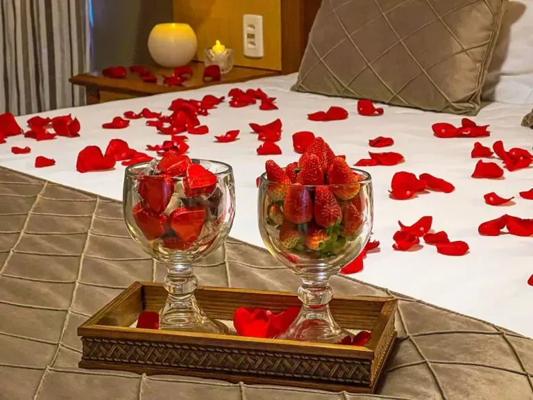 Cama decorada com pétalas de rosa, dois copos de morangos em uma bandeja.