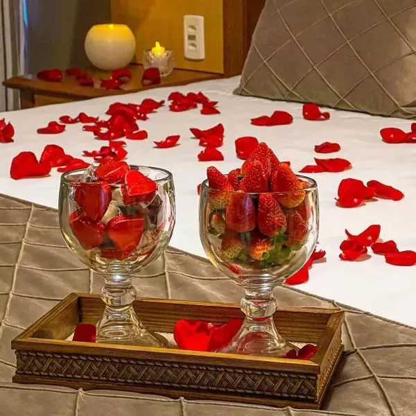 Cama decorada com pétalas de rosa, dois copos de morangos em uma bandeja.