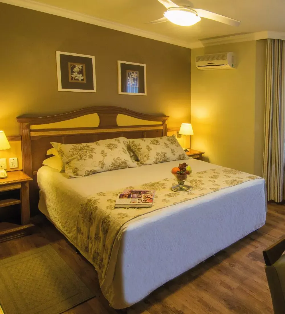 Quarto com cama king-size, roupa de cama floral, mesas de cabeceira e obras de arte emolduradas em uma parede marrom.