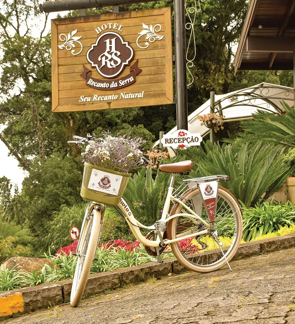 Bicicleta com cesta de flores em frente à placa do hotel "Hotel Recanto da Serra", em uma estrada inclinada.