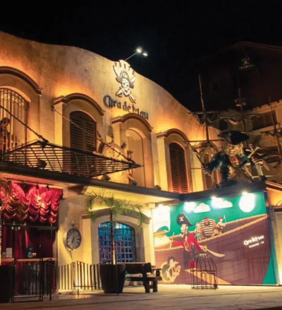 Vista noturna do edifício "O Barco do Boco". Exterior iluminado com um mural de piratas, toldo e fachada decorativa com um logotipo.