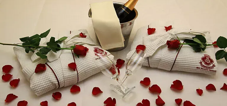 Ambiente romântico de hotel com champanhe, roupões de banho, pétalas de rosa e flores.