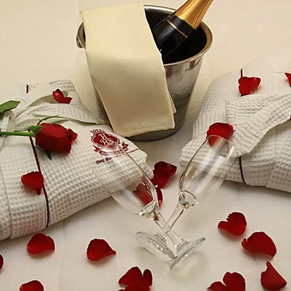 Balde de champanhe, taças, pétalas de rosa e roupões sobre uma cama. Ambiente romântico.