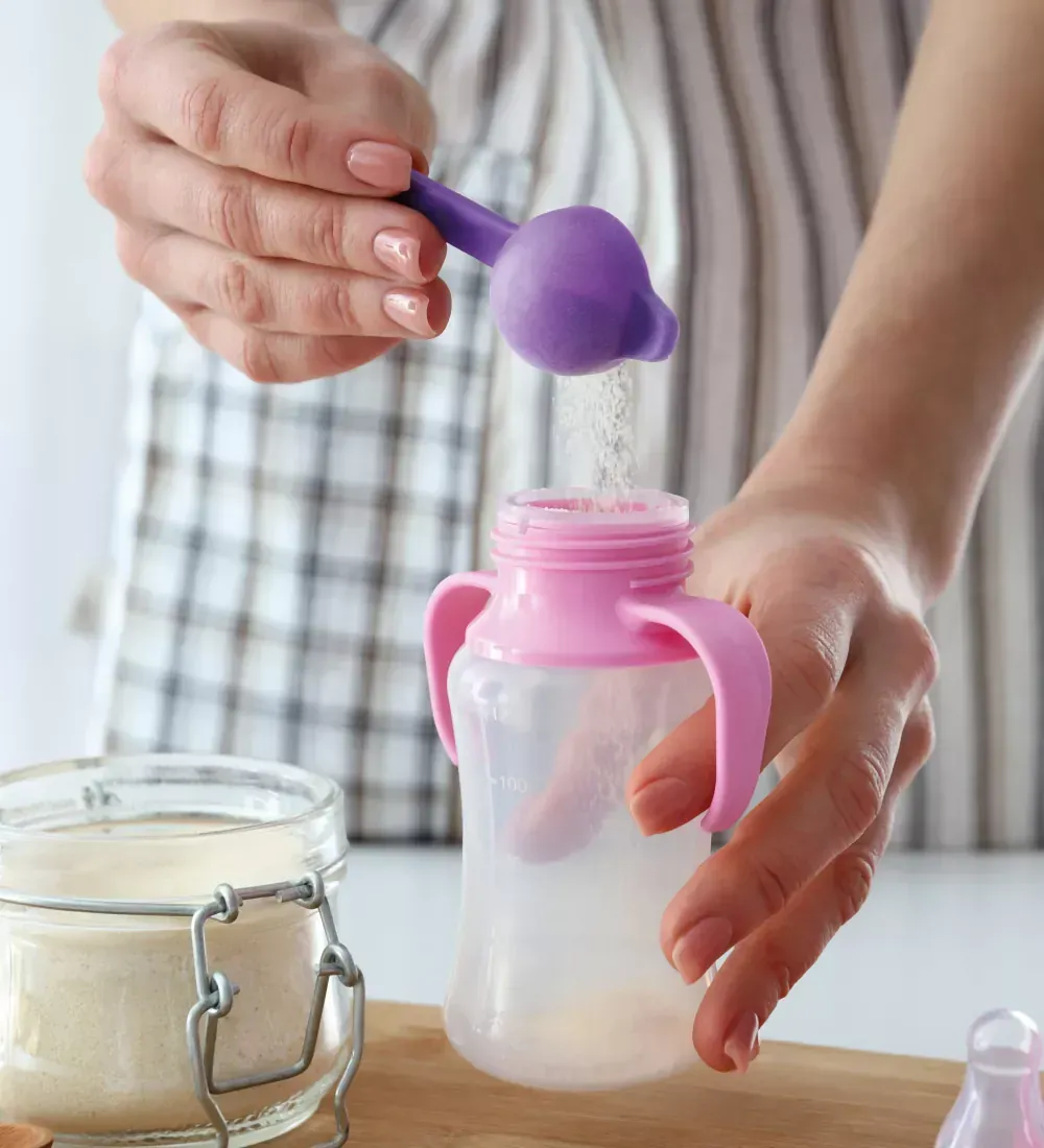 Pessoa despejando fórmula infantil em pó em uma mamadeira com alças rosas, usando uma colher medidora roxa.