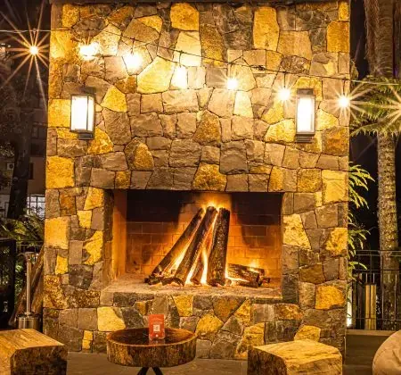 Lareira de pedra com lenha queimando, área externa com mesas e cadeiras, iluminada por apliques de parede e com assentos feitos de troncos de árvore.