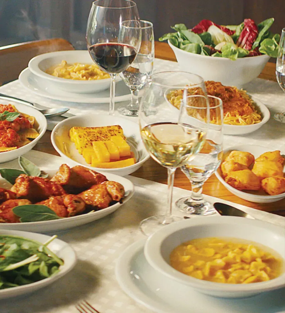 Mesa posta com diversos pratos, taças de vinho e salada. Iluminação aconchegante, louça branca.