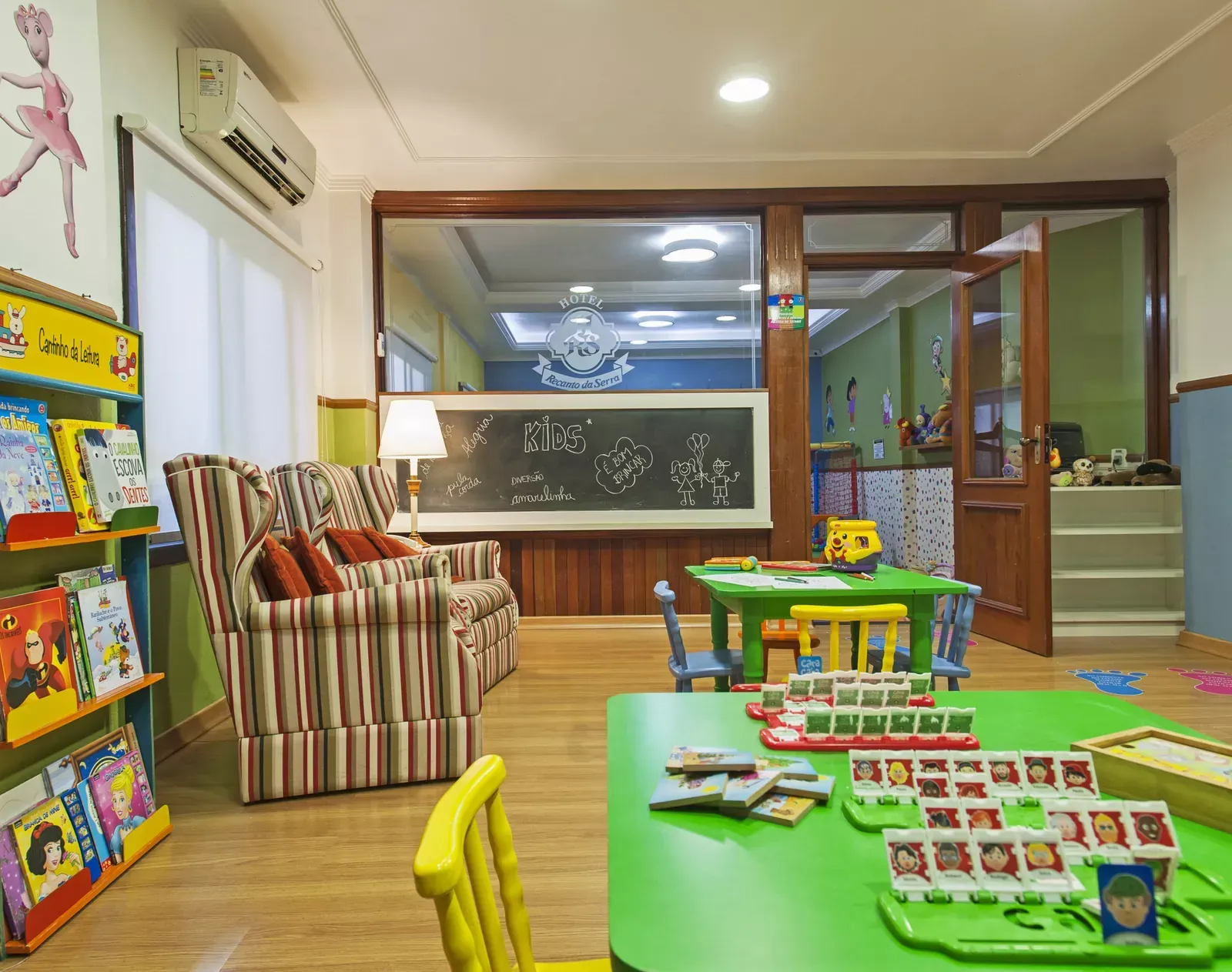 Sala de aula do jardim de infância com mesas, cadeiras, estantes de livros e área de estar verdes.