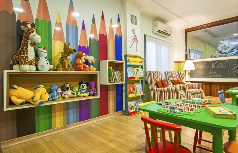Sala de jogos infantil com mural colorido de lápis na parede, brinquedos em prateleiras, mesinhas verdes e área de leitura.