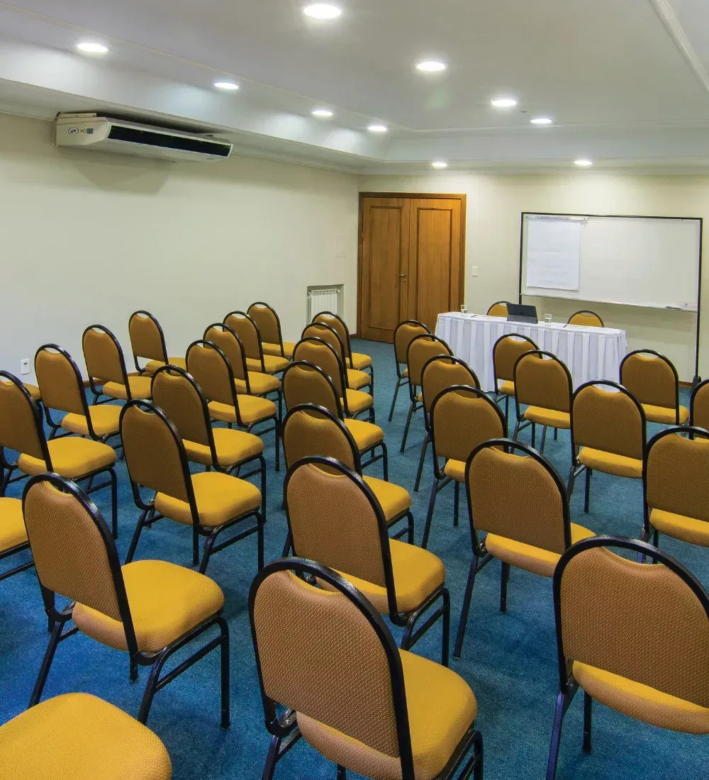 Sala de conferências vazia com fileiras de cadeiras amarelas, uma mesa e um quadro branco.