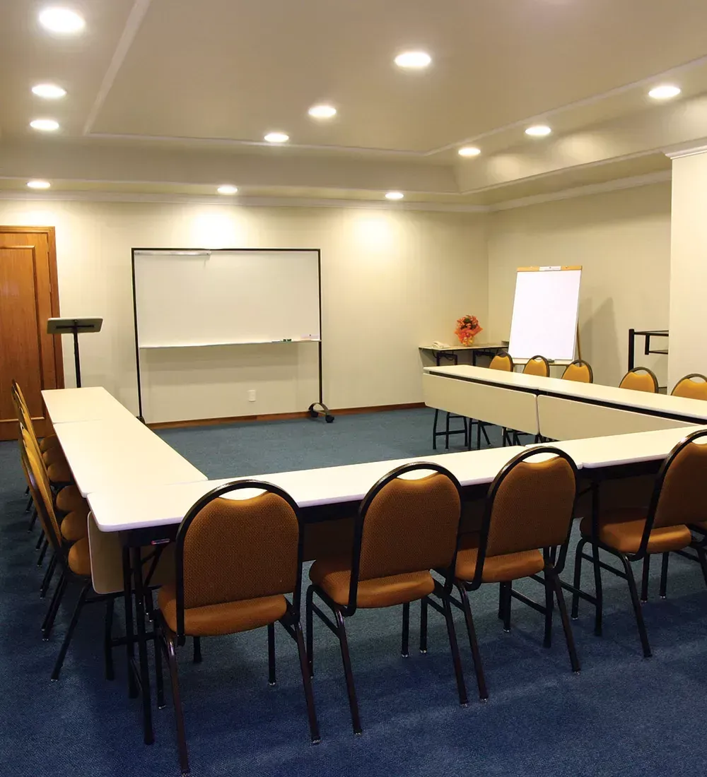 Sala de conferências com mesa e cadeiras em formato de U, quadro branco e flip chart.