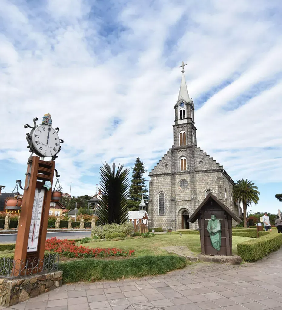 Igreja com alta torre, torre do relógio e pequena estátua em um jardim sob um céu nublado.