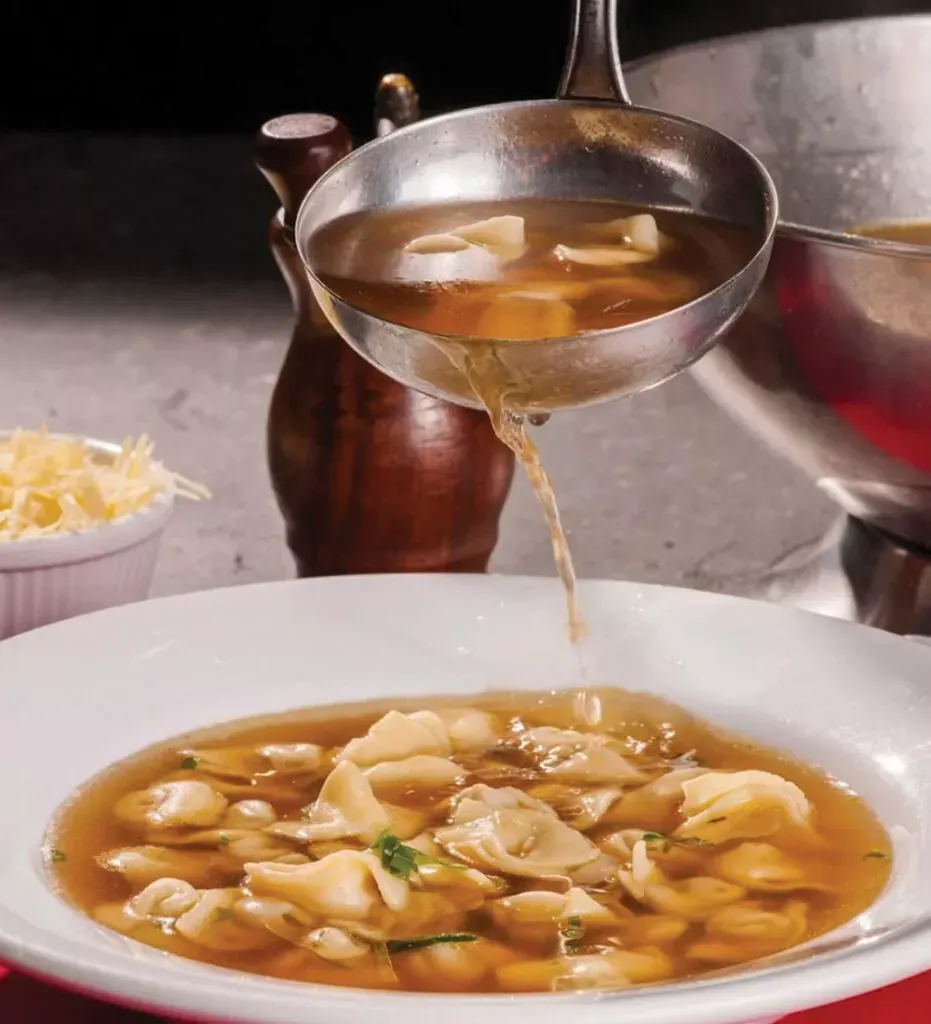 Sopa sendo servida em uma tigela branca cheia de tortellini, ao lado de uma tigela de queijo ralado.