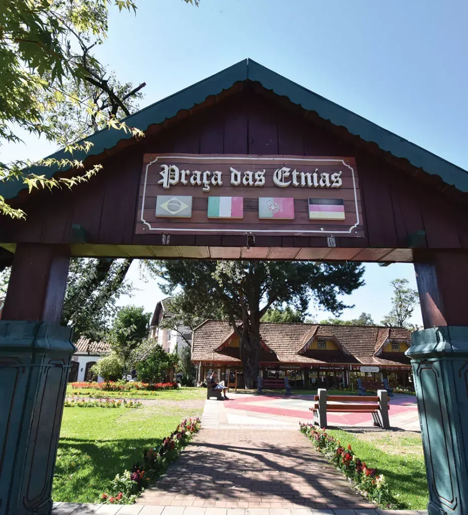 Entrada da Praça das Etnias em Gramado, Brasil, com bandeiras e um arco de madeira que dá acesso a um parque.