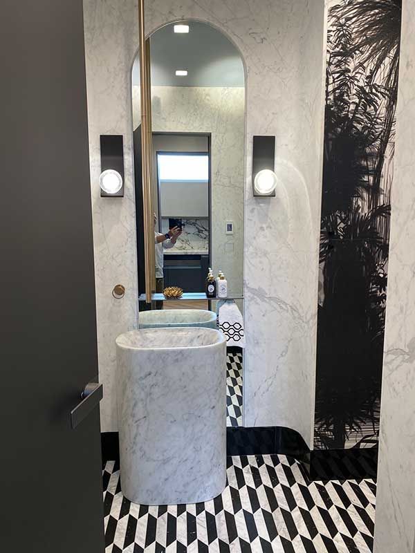 A modern powder room with a pedestal sink, black and white patterned floor, and an arched mirror.