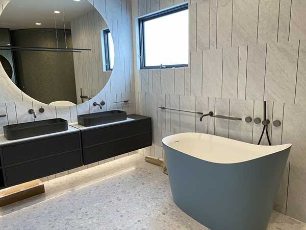 Modern bathroom with a blue tub, floating vanity, round mirror, and light-colored stone tile.
