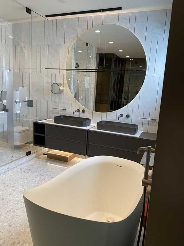 Modern bathroom with a large round mirror, double sinks, freestanding tub, and textured tile walls.
