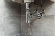 Leaking bathroom sink drain pipe against gray tiled wall. Water droplets visible.