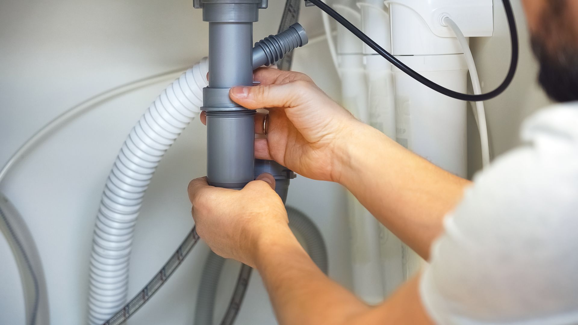 Person fixing a sink's plumbing with gray pipes. White walls are in the background.